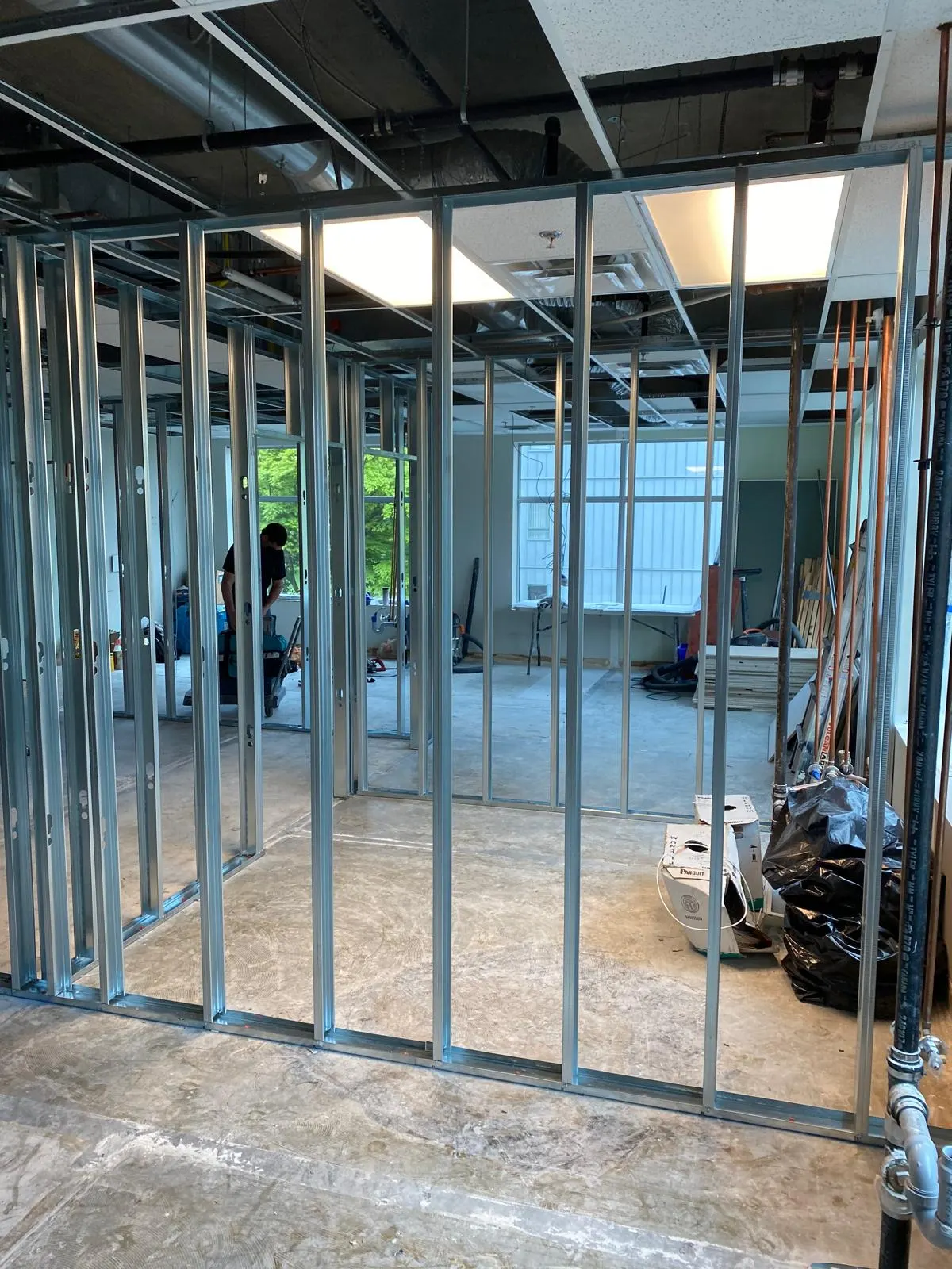 Wide view of the same office framing in progress, a worker installing hardware in a doorway with green trees visible through the window