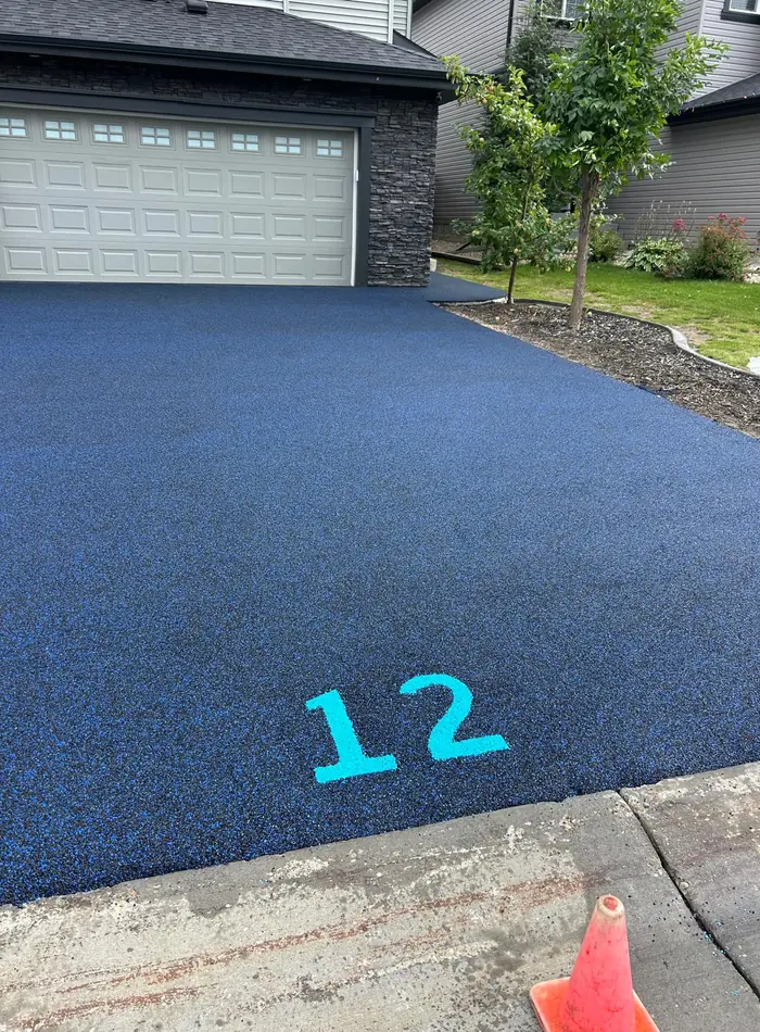 Residential driveway surfaced in blue-flecked poured rubber with a house number inlaid in bright turquoise, running up to a two-car garage